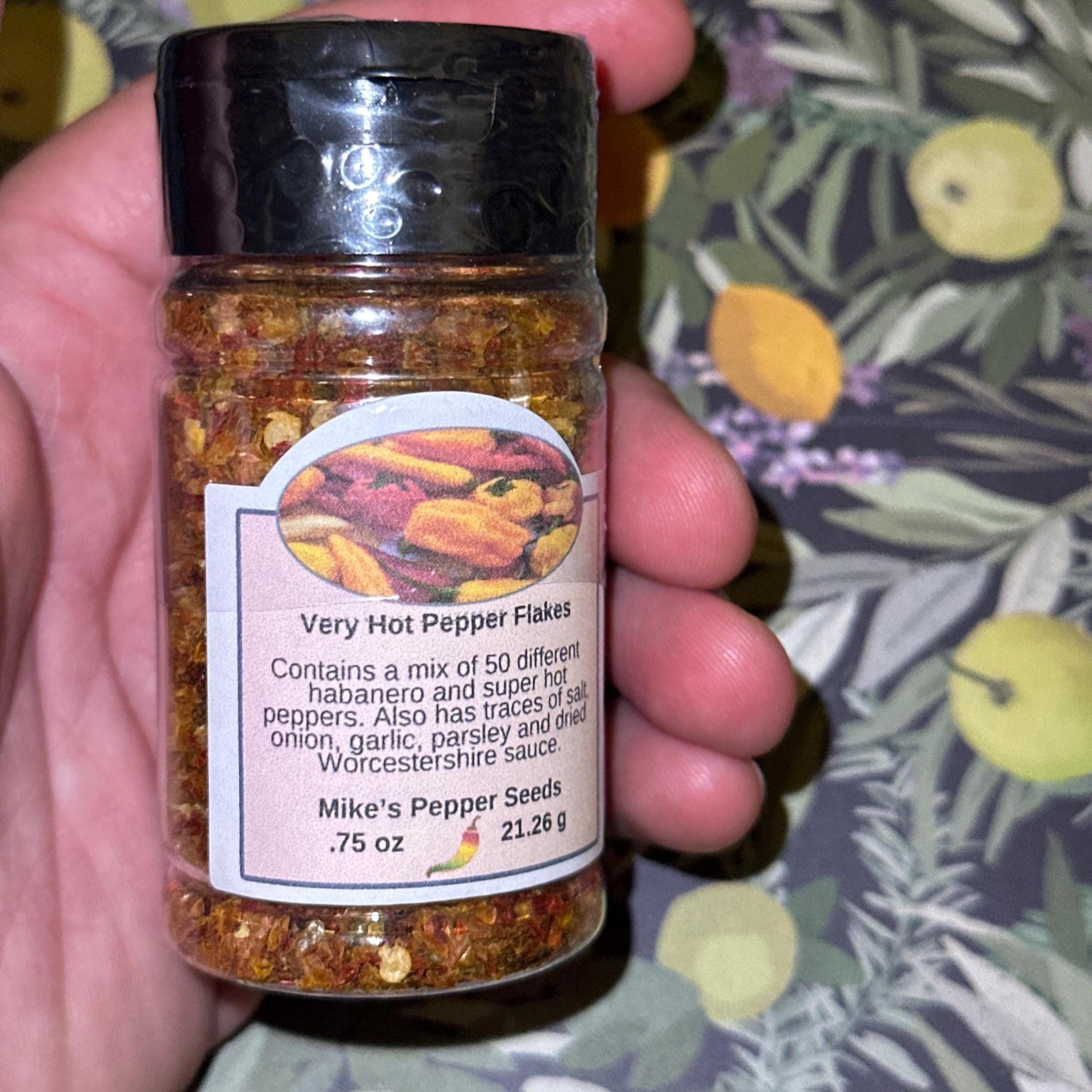 Mike’s Pepper Seeds Hand holding a jar of very hot pepper flakes with a colorful label against a floral-patterned background.